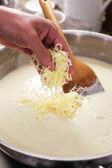 A person dropping a handful of finely shredded fontina cheese in a skillet filled with a creamy bechamel sauce.