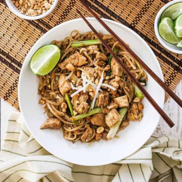 An overhead view of a white bowl filled with homemade chicken pad Thai with a pair of chopsticks on the side of the bowl.
