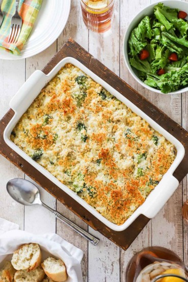 An overhead view of a 9 by 13 white baking dish filled with a chicken and rice casserole topped with toasted breadcrumbs and flanked with a bowl of a green leaf salad and a basket of bread pieces.