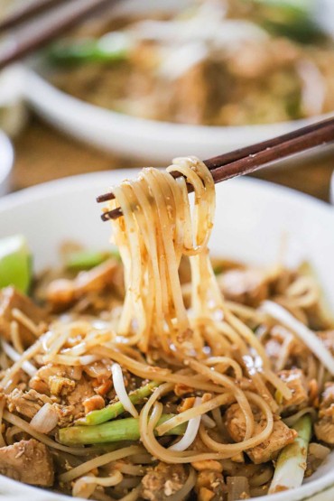 A pair of chopsticks lifting rice noodles from a bowl filled with chicken pad Thai.