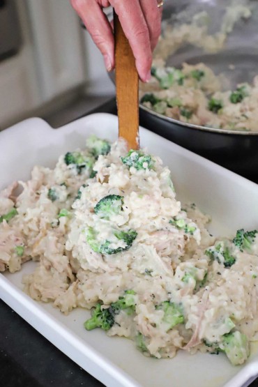 A person using a wooden spatula to transfer the contents of chicken and rice casserole into a 9 by 13 white baking dish.