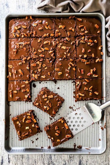 An overhead view of a sheet pan half filled with Texas sheet cake that has been cut into squares with a metal spatula holding one of the square near the edge of the pan.