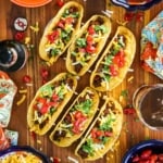 Six Tex-Mex Beef Tacos sitting up-right in two taco stands sitting on a cutting board surrounded by bowls of traditional toppings.