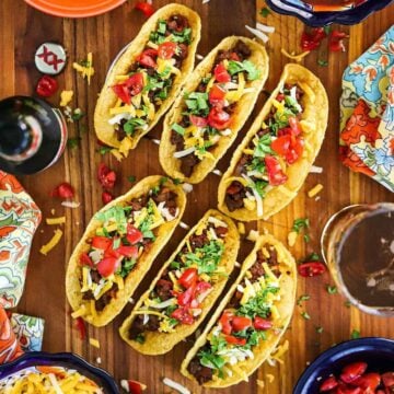 Six Tex-Mex Beef Tacos sitting up-right in two taco stands sitting on a cutting board surrounded by bowls of traditional toppings.