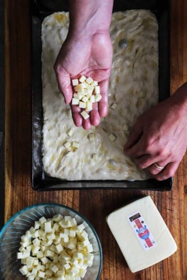 An overhead view of a person holding small cubes of brick cheese with one hand and placing those cubes into pizza dough in a Detroit-style pizza pan.