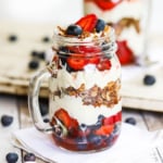 A straight-on view of a glass jar with a handle holding a wild berry parfait with mascarpone and topped with strawberries, blueberries, and granola.