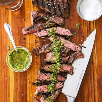 An overhead view of a sous vide skirt steak on a cutting board with half of it cut into strips and topped with homemade chimichurri sauce.