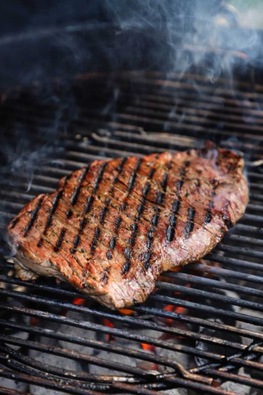 A whole skirt steak searing on a grill gate over a hot and smoking pile of ashy charcoal briquettes.