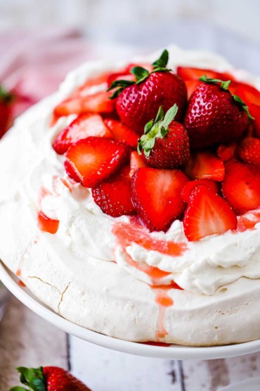 An untouched strawberry pavlova topped with macerated strawberries and fresh strawberries sitting on a white cake stand.