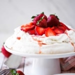 A side view of a strawberry pavlova sitting on a white cake stand next to a red-checkered napkin.