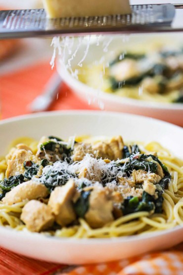 A close-up view of a white pasta bowl filled with creamy chicken florentine over pasta with a person grating parmesan cheese over the top. 