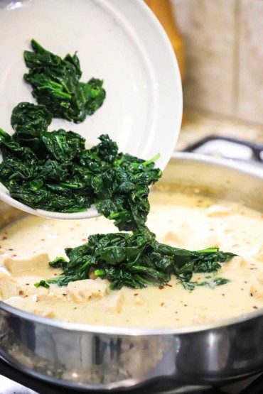 A person dumping sautéed spinach from a white bowl into a skillet filled with cooked chicken chunks in a creamy sauce. 