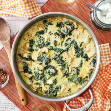 An overhead view of a large silver skillet filled with creamy chicken Florentine with a wooden spoon and small bowl of Parmesan cheese sitting nearby.