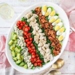 An overhead view of a Cobb salad on a large white oval platter flanked by two glasses of white wine and two gold serving spoons.