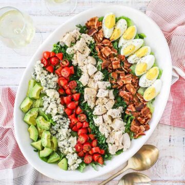 An overhead view of a Cobb salad on a large white oval platter flanked by two glasses of white wine and two gold serving spoons.