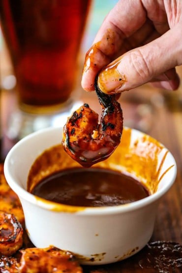 A person lifting a grilled BBQ shrimp out a small bowl filled with homemade BBQ sauce.