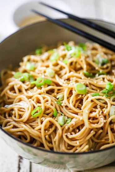 A close-up view of an Asian-style bowl filled with Chinese noodles with peanut sauce and topped with chopped scallions and sesame seeds.