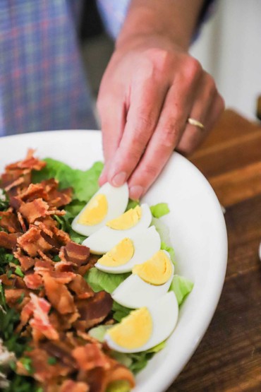 A person placing a hard boiled egg that has been quartered onto an oval platter with a row of other eggs which are arranged next to a row of crumbled cooked bacon.