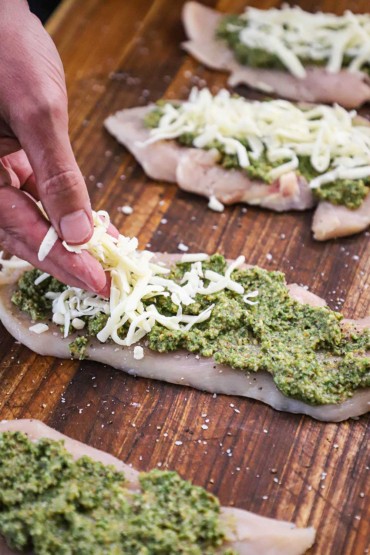A person sprinkling shredded mozzarella cheese over the top of a thin uncooked chicken cutlet that has a layer of pesto on it. 