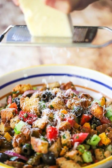 A person using a microplane to grate a block of Parmesan cheese over a bowl filled with panzanella (bread and tomato salad).