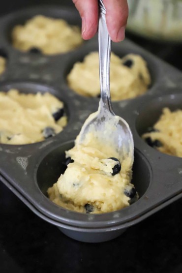 A person using a large silver spoon to transfer lemon and blueberry muffin batter into a jumbo muffin pan.