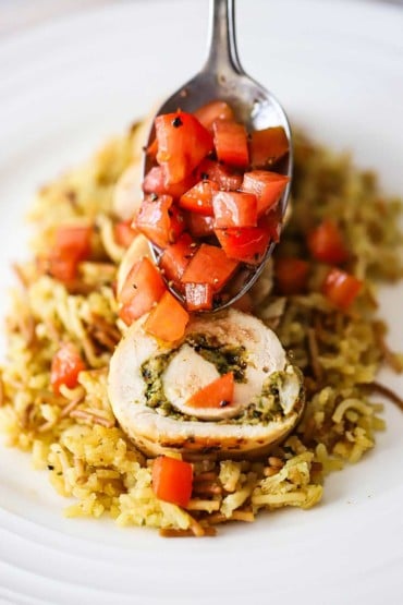 A person using a spoon to transfer a chopped tomato relish over slice pieces of cooked rolled up chicken that has been stuffed with pesto sauce.