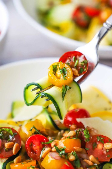 A person using a fork to hold up a softened strip of zucchini that is topped with tomato relish over a bowl of the same.