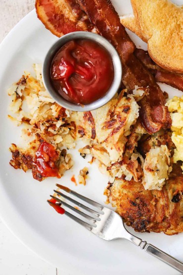 An overhead view of a plate filled with a half-eaten portion of hash browns with a small container of ketchup, crisp bacon, and sliced toast nearby