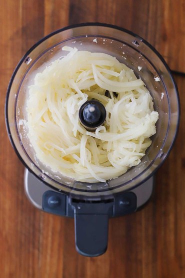 An overhead view of a food processor that is filled with shredded par-boiled russet potatoes.