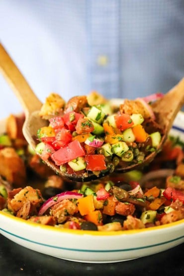 A person using two large wooden spoons to toss together chopped vegetables and homemade croutons in a large pasta bowl.