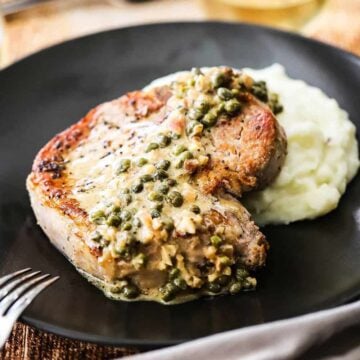 A front-on view of a black plate filled with mashed potatoes and a seared pork chop with caper sauce on top of it.