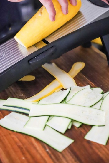 A person using a mandolin slicer to cut a yellow squash into long thin strips that resemble sheets of yellow pasta.