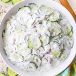 An overhead view of a white bowl filled with Tzatziki Cucumber Salad that is flanked by a wooden spoon and freshly sliced cucumbers.