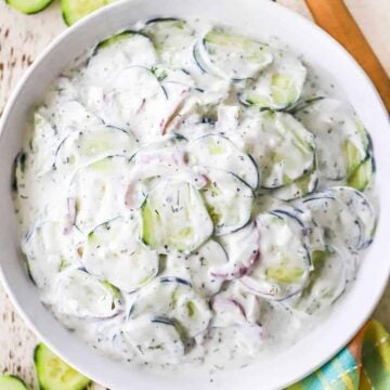 An overhead view of a white bowl filled with Tzatziki Cucumber Salad that is flanked by a wooden spoon and freshly sliced cucumbers.
