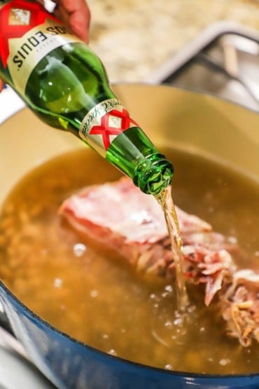 A person pouring a beer into a large Dutch oven filled with chicken stock and a ham bone.