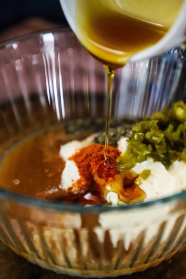 Dark corn syrup being poured from a small white bowl into a larger glass bowl filled with the ingredients for Alabama white sauce.