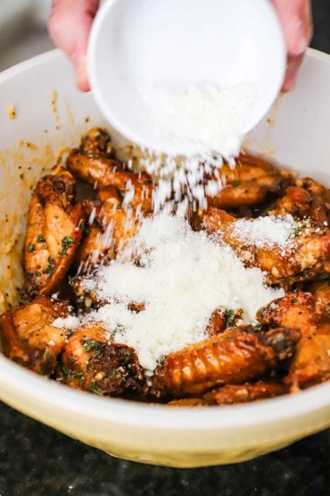 A person dumping a small bowl filled with grated Parmesan cheese into a ceramic bowl that is filled with cooked and sauced chicken wings.