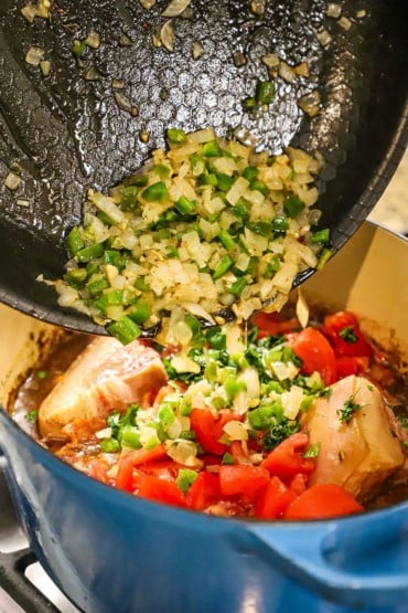 A person dumping sautéed onions and jalapeños into pot filled with chopped tomatoes, a hambone, and chicken stock.