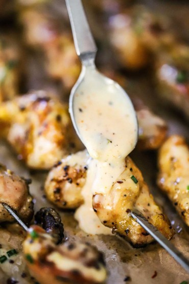 Alabama white sauce transferring onto a piece of skewered grilled chicken from a large silver spoon.