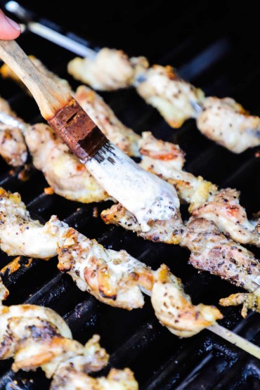 A person using a small kitchen brush to apply Alabama white sauce over skewered pieces of chicken that are on the grates of a gas grill.