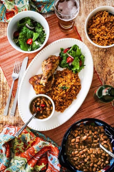 An overhead view of a white platter filled with a serving of Mexican rice, grilled chicken, and a bowl of borracho beans surrounded by bowls of the same.