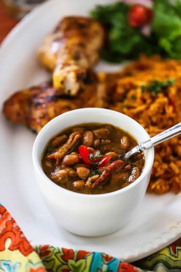 A close-up view of a white ceramic cup filled with a serving of borracho beans on a platter surrounded by rice and grilled chicken.