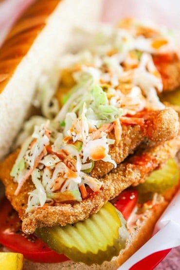 A close-up view of a fried catfish po-boy with homemade coleslaw and hot sauce sitting on top of the catfish, pickles, and sliced tomatoes, all on a soft roll.