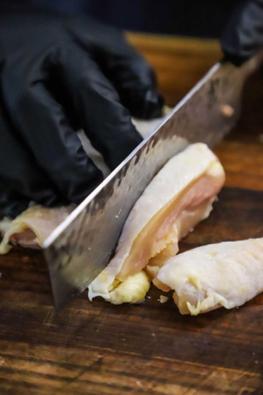A person wearing black latex gloves using a large Japanese knife to cut strips of a chicken thigh from the bone.