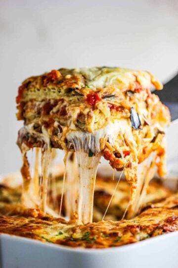 A square slice of baked eggplant parmesan being lifted from the dish with mozzarella strands hanging from the spatula.