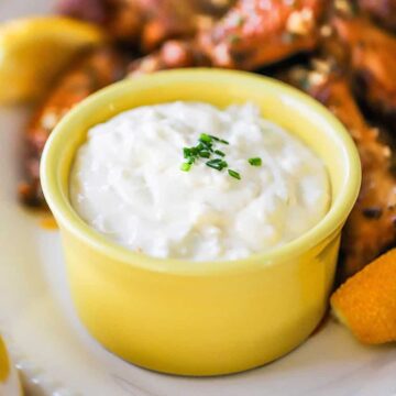 A straight-on view of a small yellow bowl that is filled with a gorgonzola dipping sauce and is sitting on a platter next to a pile of cooked wings.