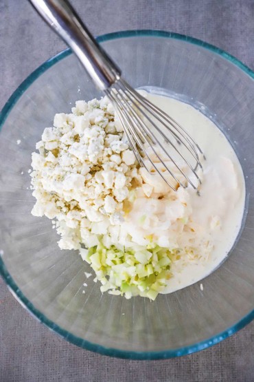 An overhead view of a medium-sized glass bowl holding crumbled gorgonzola cheese, diced celery and onion, mayonnaise, and cream.