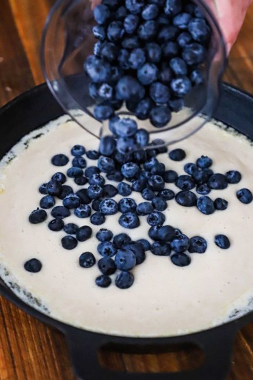 A person dumping fresh blueberries from a glass measuring cup into a cast-iron skillet filled with uncooked cobbler batter.