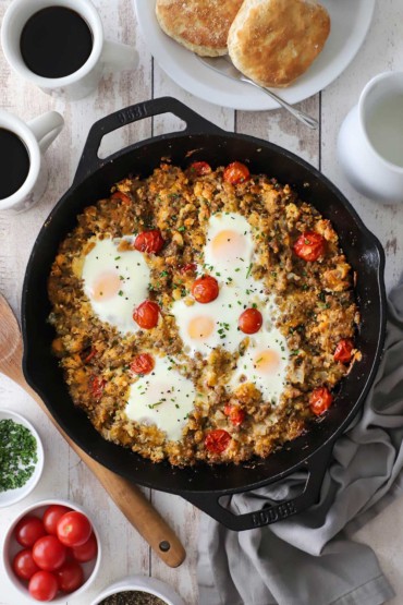 An overhead view of a skillet breakfast of cooked sausage mixed with crumbled biscuits, cherry tomatoes, and cheese all topped with five cooked eggs.