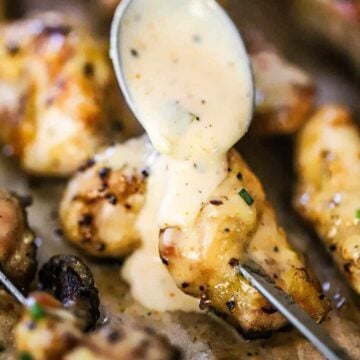 Alabama white sauce transferring onto a piece of skewered grilled chicken from a large silver spoon.